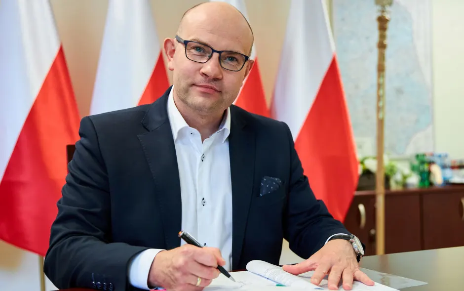 Na zdjęciu Artur Kosicki Marszałek województwa Podlaskiego siedząc za biurkiem podpisuje dokumenty. W tle polskie flagi.