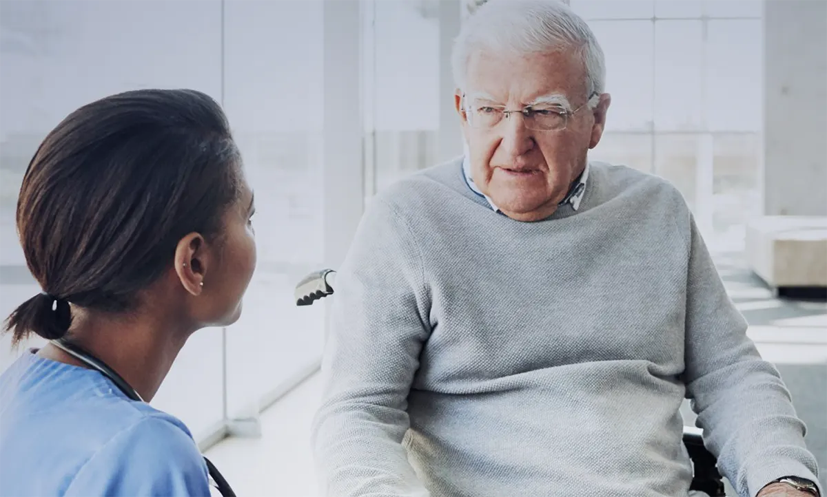 Mężczyzna na wózku, spogląda w kierunku kobiety, prawdopodobnie pielęgniarki